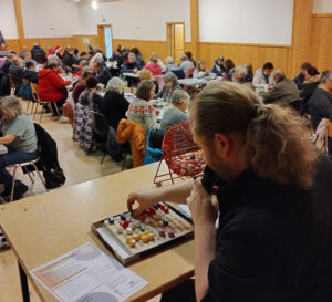 Bingokoneen pyörittäjä Joni laittaa bingopalloa alustalle, taustalla bingon pelaajia.